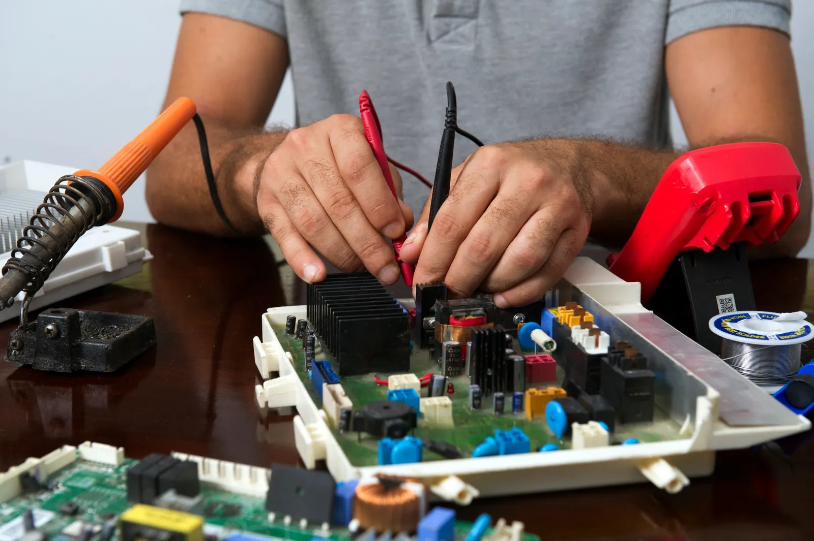 Technician testing a circuit board with a multimeter and soldering iron during a board-level electronics repair